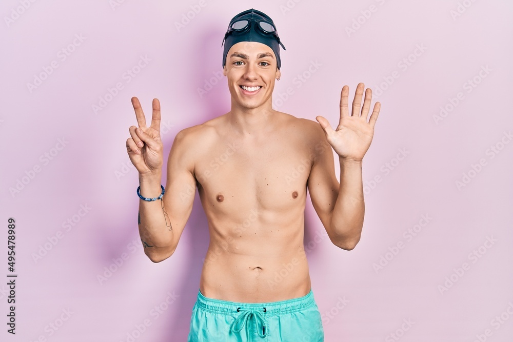Young hispanic man wearing swimwear and swimmer glasses showing and pointing up with fingers number seven while smiling confident and happy.