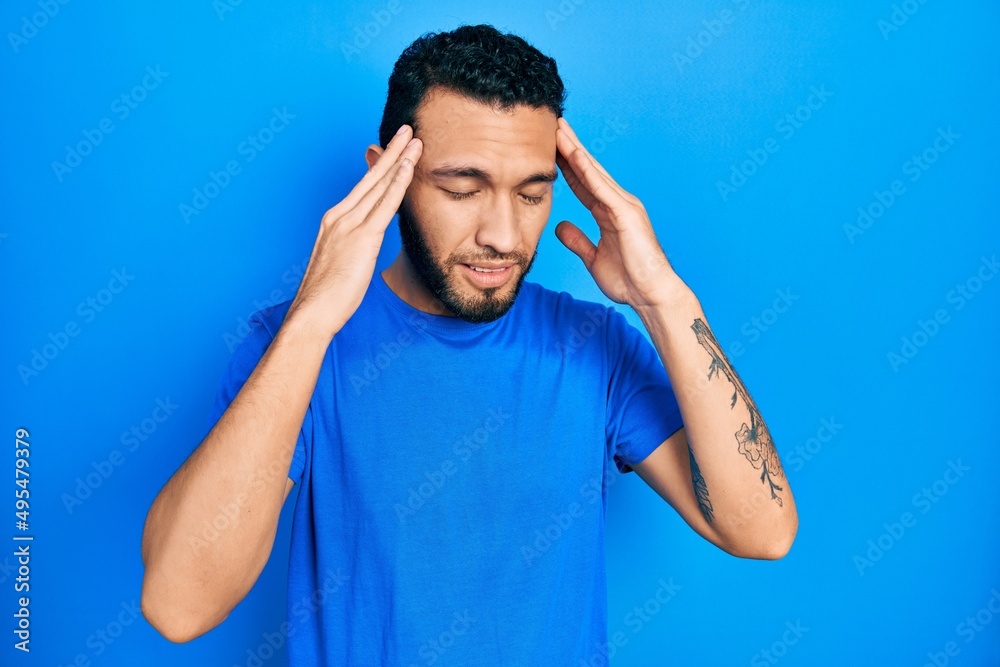 Hispanic man with beard wearing casual blue t shirt with hand on head for pain in head because stress. suffering migraine. Hispanic man with beard wearing casual blue t shirt with hand on head for pain in head because stress. suffering migraine.