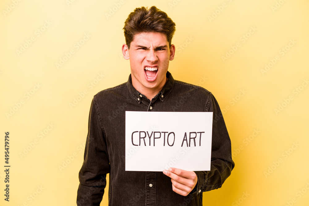 Young caucasian man holding crypto art placard isolated on yellow background screaming very angry and aggressive. Young caucasian man holding crypto art placard isolated on yellow background screaming very angry and aggressive.
