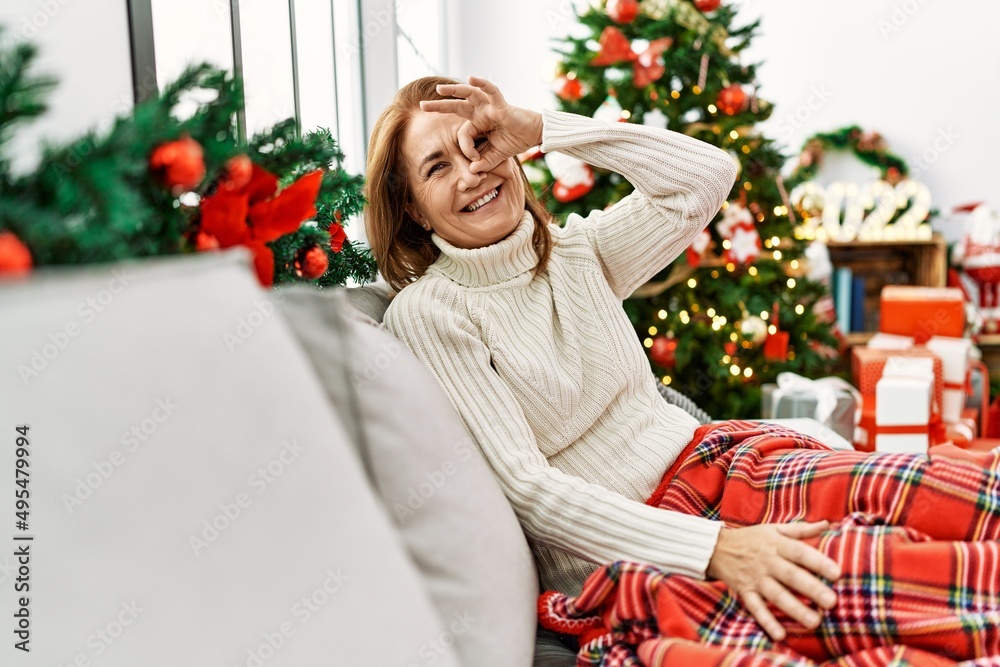 Middle age woman sitting on the sofa by christmas tree smiling happy doing ok sign with hand on eye looking through fingers Middle age woman sitting on the sofa by christmas tree smiling happy doing ok sign with hand on eye looking through fingers