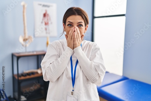 Young brunette woman working at pain recovery clinic laughing and embarrassed giggle covering mouth with hands, gossip and scandal concept © Krakenimages.com