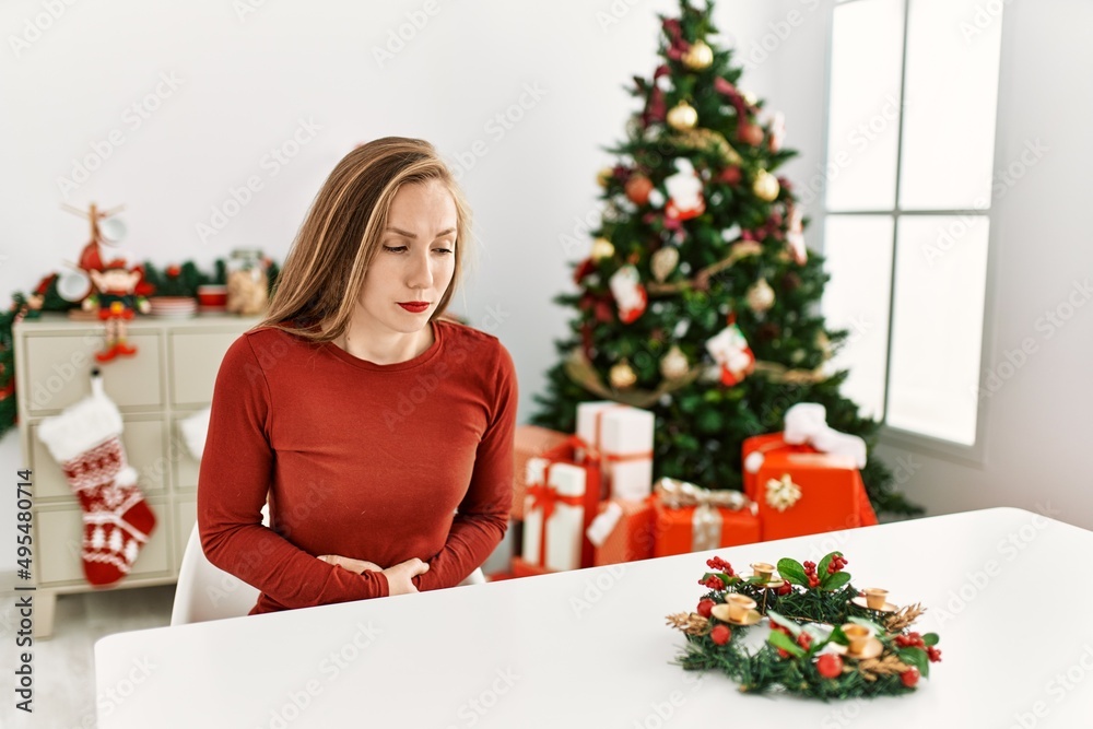 Caucasian young blonde woman sitting on the table by christmas tree with hand on stomach because nausea, painful disease feeling unwell. ache concept. Caucasian young blonde woman sitting on the table by christmas tree with hand on stomach because nausea, painful disease feeling unwell. ache concept.