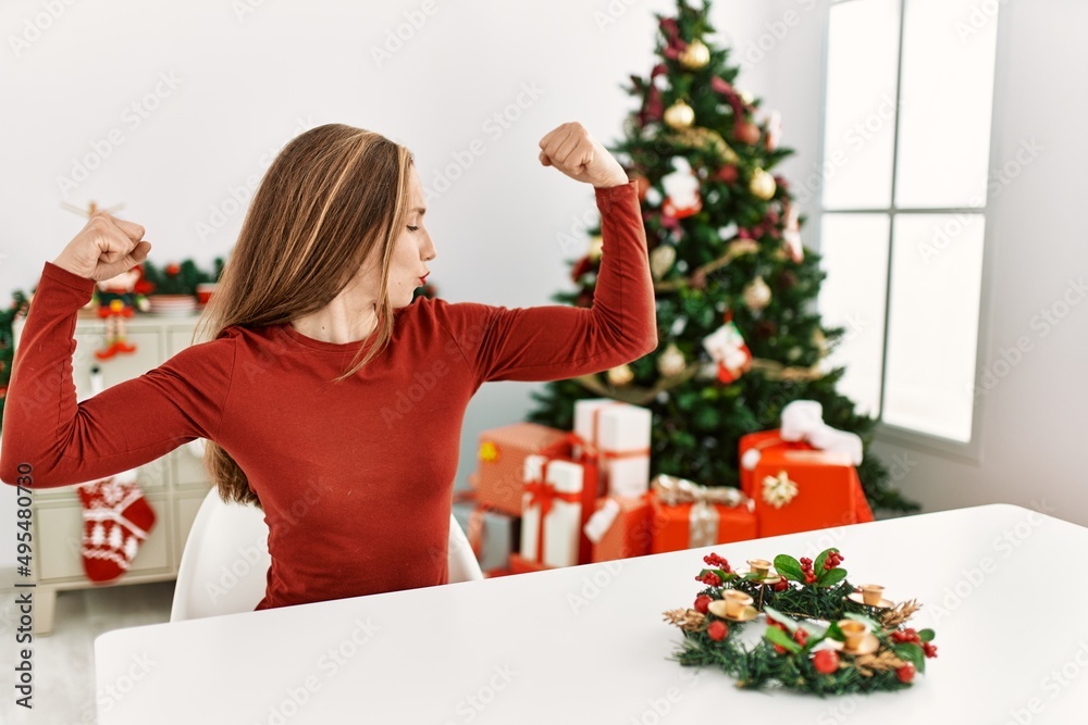 Caucasian young blonde woman sitting on the table by christmas tree showing arms muscles smiling proud. fitness concept. Caucasian young blonde woman sitting on the table by christmas tree showing arms muscles smiling proud. fitness concept.