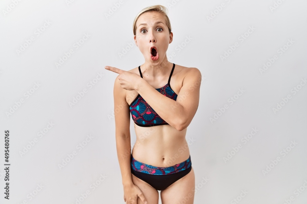Young blonde woman wearing sporty bikini over isolated background surprised pointing with finger to the side, open mouth amazed expression. Young blonde woman wearing sporty bikini over isolated background surprised pointing with finger to the side, open mouth amazed expression.