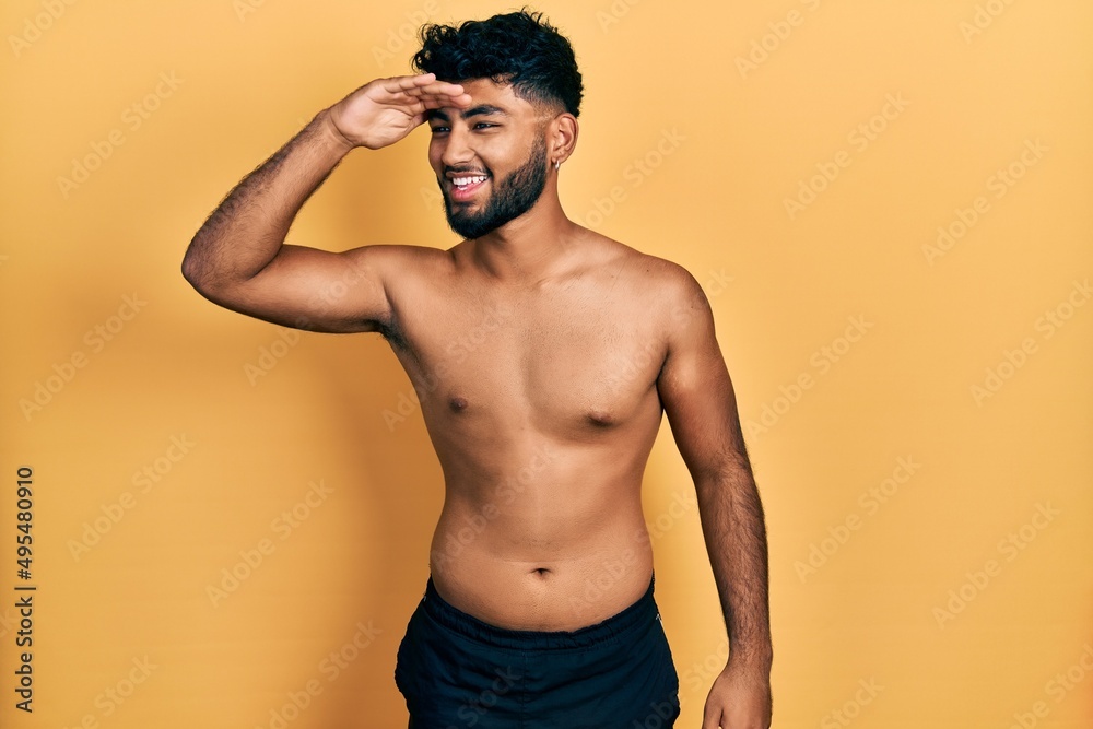 Arab man with beard wearing swimwear shirtless very happy and smiling looking far away with hand over head. searching concept.