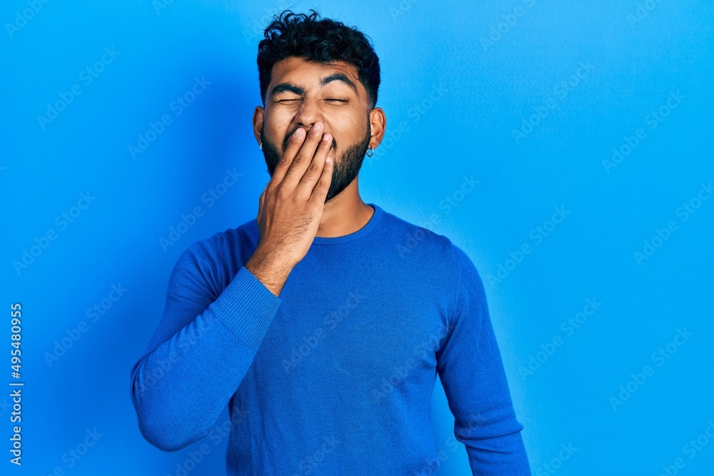 Arab man with beard wearing casual blue sweater bored yawning tired covering mouth with hand. restless and sleepiness.