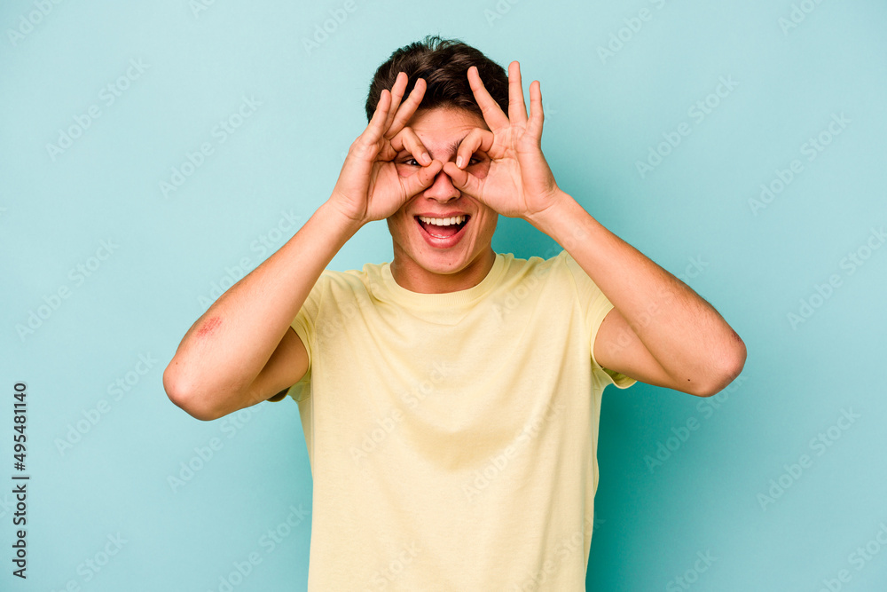 Young caucasian man isolated on blue background showing okay sign over eyes Young caucasian man isolated on blue background showing okay sign over eyes