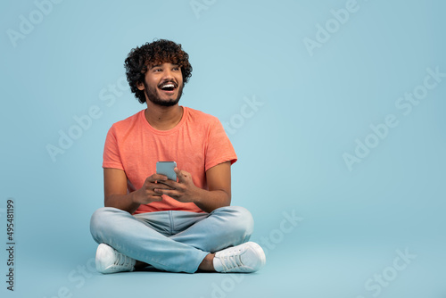 Excited indian guy sitting on floor on blue, using smartphone © Prostock-studio