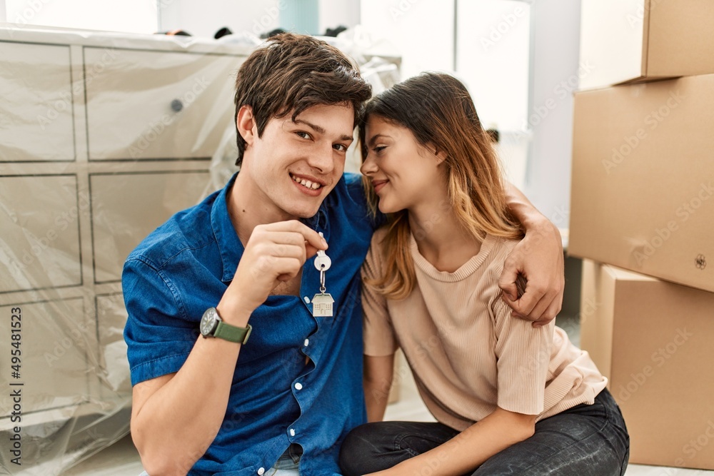 Young caucasian couple smiling happy sitting on the floor holding key of new home.