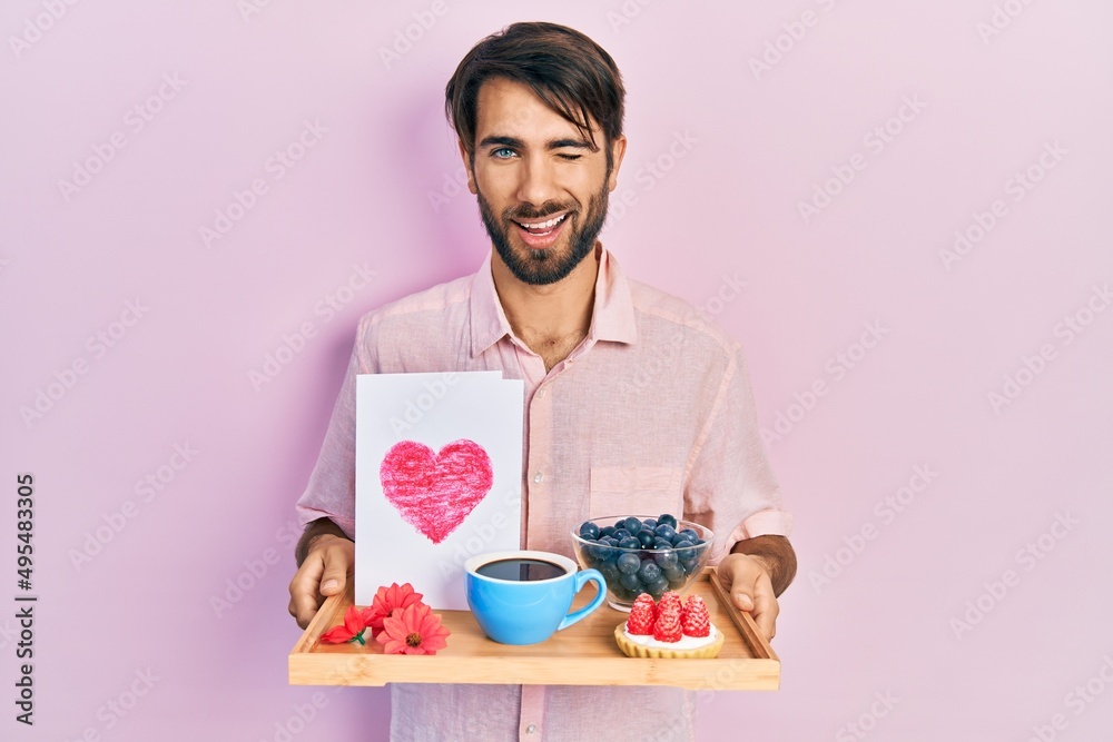 Young hispanic man holding tray with breakfast food and heart draw winking looking at the camera with sexy expression, cheerful and happy face. Young hispanic man holding tray with breakfast food and heart draw winking looking at the camera with sexy expression, cheerful and happy face.