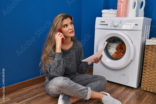 Young blonde woman talking on the smartphone waiting for washing machine at laundry room