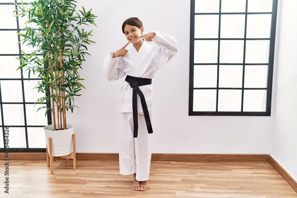 Young hispanic girl wearing karate kimono and black belt smiling cheerful showing and pointing with fingers teeth and mouth. dental health concept. Young hispanic girl wearing karate kimono and black belt smiling cheerful showing and pointing with fingers teeth and mouth. dental health concept.
