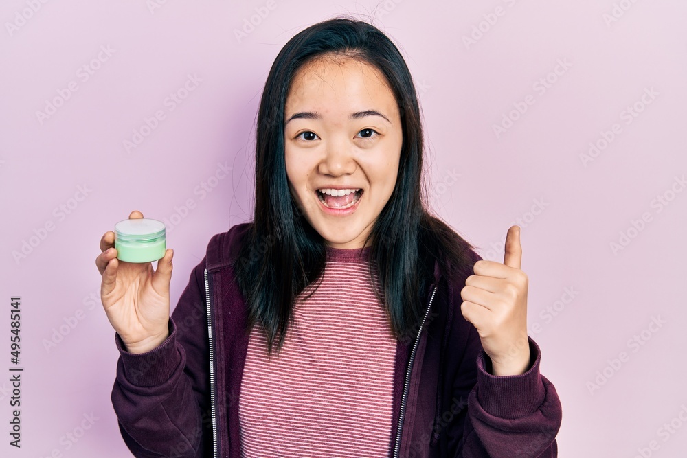 Young chinese girl holding cosmetic moisturizer facial cream screaming proud, celebrating victory and success very excited with raised arms Young chinese girl holding cosmetic moisturizer facial cream screaming proud, celebrating victory and success very excited with raised arms