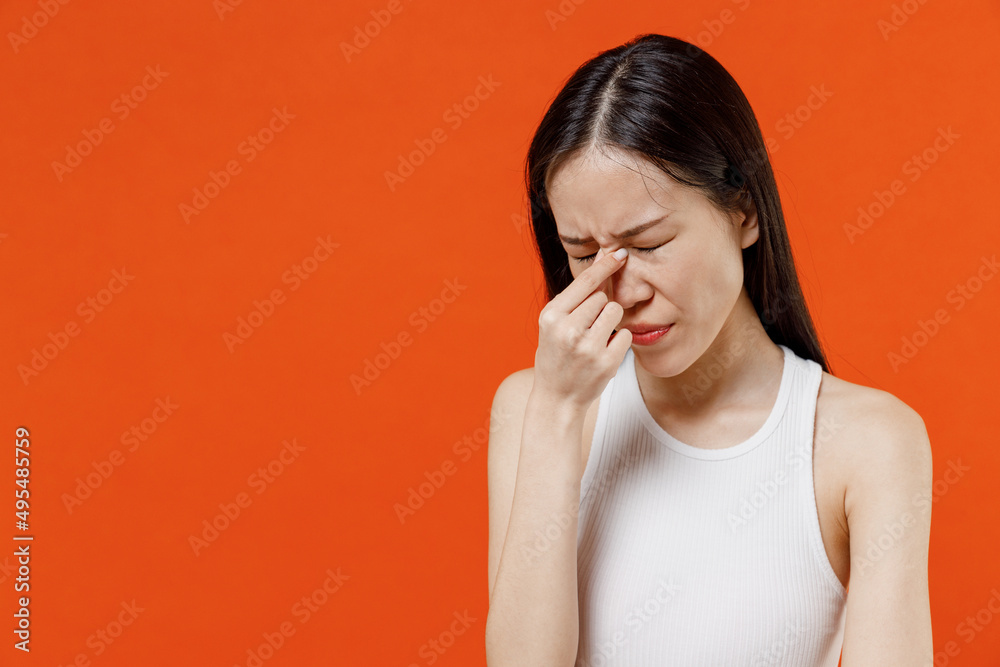 Frustrated tired exhausted unhappy preoccupied young woman of Asian ethnicity 20s years old in white tank top keep eyes closed rub put hand on nose isolated on plain orange background studio portrait. Frustrated tired exhausted unhappy preoccupied young woman of Asian ethnicity 20s years old in white tank top keep eyes closed rub put hand on nose isolated on plain orange background studio portrait.