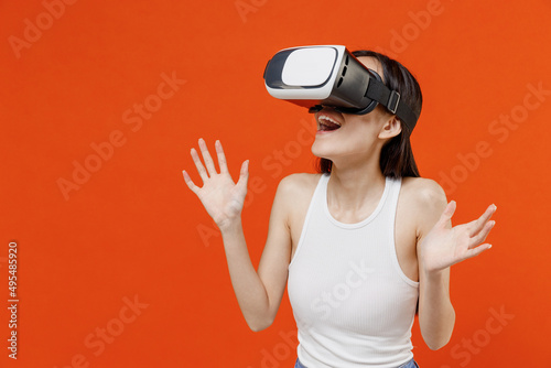 Shocked surprised fun cheerful blithesome stupefied young woman of Asian ethnicity 20s years old in white tank top watching in vr headset pc gadget isolated on plain orange background studio portrait. © ViDi Studio