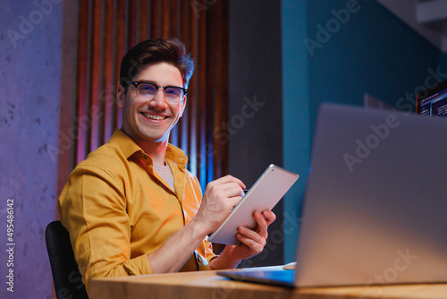 Young happy professional fun content creator IT specialist programmer man in shirt work at office on laptop pc computer using tablet searching information browse internet. Program development concept. photo