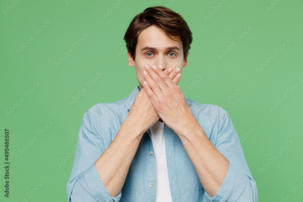 Frightened afraid scared shocked surprised young brunet man 20s years old wears blue shirt cover mouth with hand isolated on plain green background studio portrait. People emotions lifestyle concept. Frightened afraid scared shocked surprised young brunet man 20s years old wears blue shirt cover mouth with hand isolated on plain green background studio portrait. People emotions lifestyle concept.