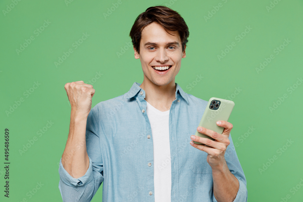 Excited fun young brunet man 20s years old wear blue shirt hold in hand use mobile cell phone doing winner gesture celebrate clenching fists say yes isolated on plain green background studio portrait Excited fun young brunet man 20s years old wear blue shirt hold in hand use mobile cell phone doing winner gesture celebrate clenching fists say yes isolated on plain green background studio portrait