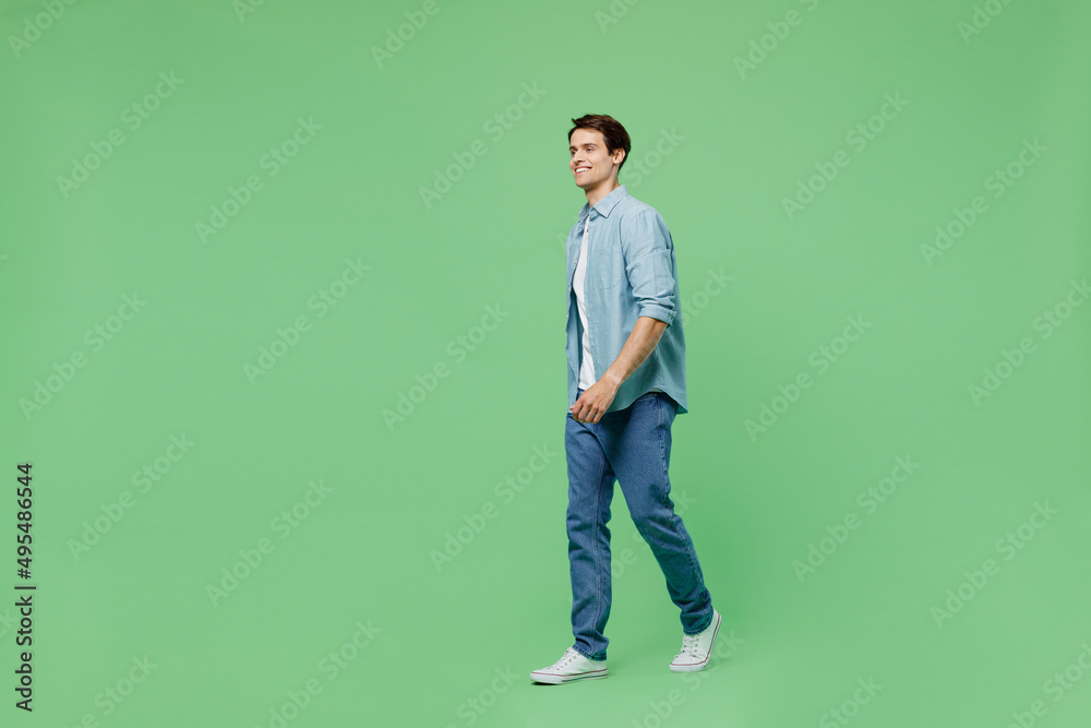 Full size body length side view blithesome charismatic stunning delight young brunet man 20s years old wears blue shirt looking aside strolling pace isolated on plain green background studio portrait. Full size body length side view blithesome charismatic stunning delight young brunet man 20s years old wears blue shirt looking aside strolling pace isolated on plain green background studio portrait.
