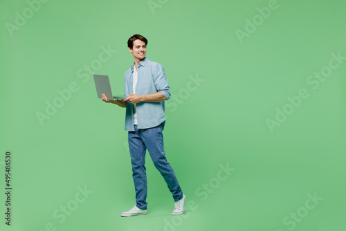 Full size body length side view smiling young brunet man 20s years old wears blue shirt hold use work on laptop pc computer looking aside step walk isolated on plain green background studio portrait.