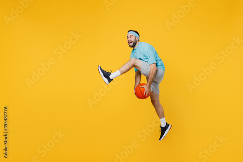 Full size caucasian young fitness trainer instructor sporty man sportsman in headband blue t-shirt hold ball play basketball game jump high isolated on plain yellow background. Workout sport concept.