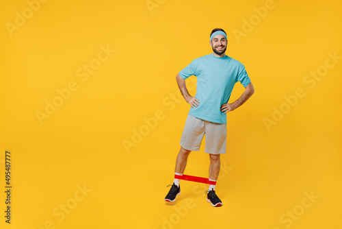 Full body young smiling happy fitness trainer instructor sporty man sportsman in headband blue t-shirt use fitness elastic bands stand akimbo isolated on plain yellow background Workout sport concept