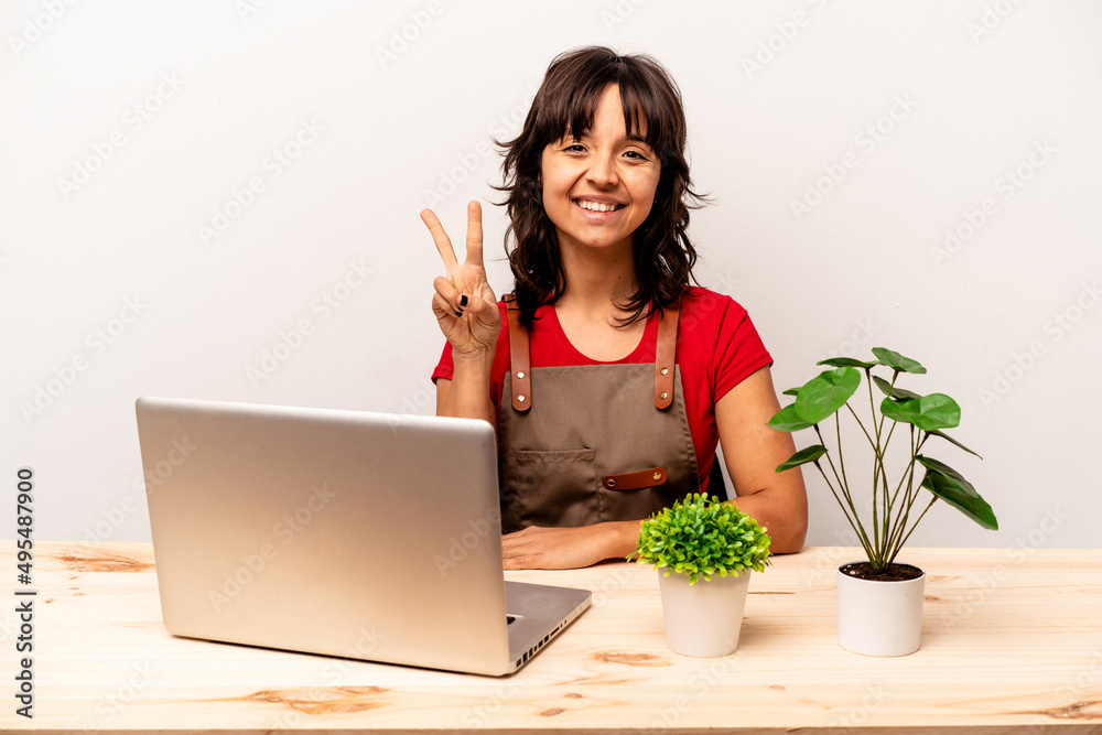Young gardener hispanic woman sitting on a table isolated on white background showing number two with fingers. Young gardener hispanic woman sitting on a table isolated on white background showing number two with fingers.