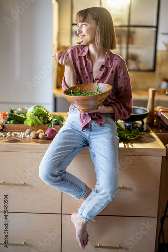 Portrait of a young cheerful woman dressed casually sitting on the table and eating healthy salad in the kitchen at home. Wellness and healthy lifestyle concept