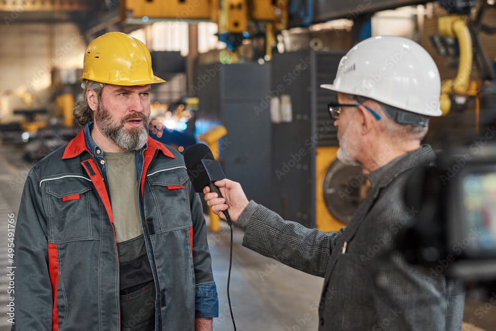 Mature worker in uniform giving an interview to microphone for reporter while they standing in workshop in factory Mature worker in uniform giving an interview to microphone for reporter while they standing in workshop in factory