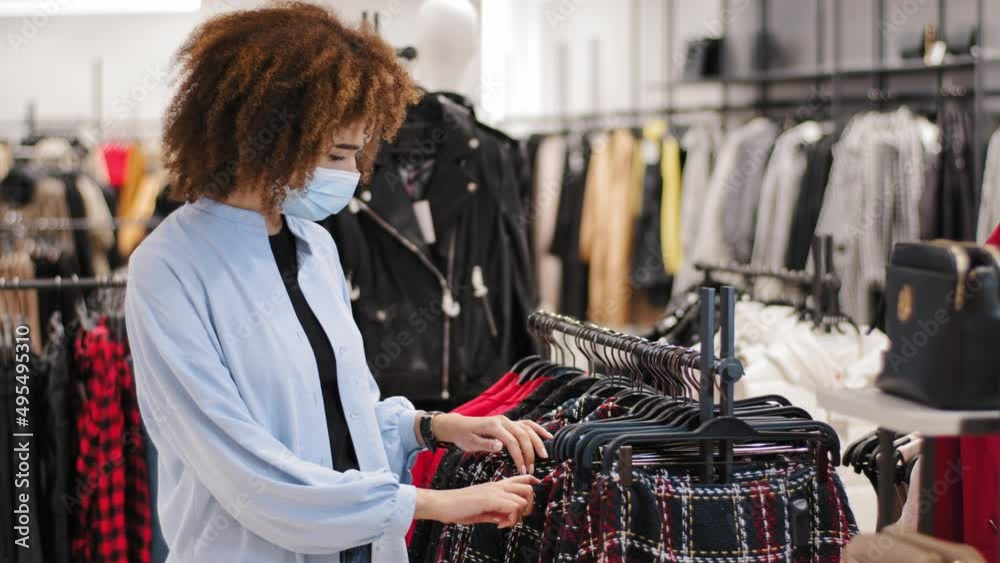 Young african woman wearing safety medical mask choosing clothes in rack in cloth shop. Multiracial curly biracial female customer buyer in protective face respirator shopping clothes in retail store Young african woman wearing safety medical mask choosing clothes in rack in cloth shop. Multiracial curly biracial female customer buyer in protective face respirator shopping clothes in retail store