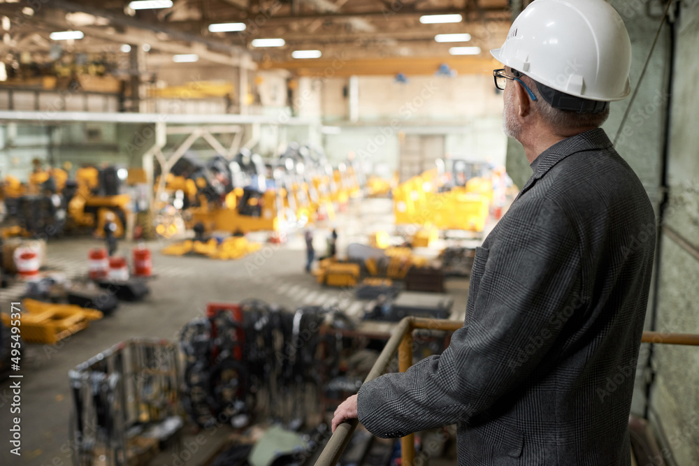 Rear view of mature engineer in work helmet standing on balcony and watching for the manufacturing in plant