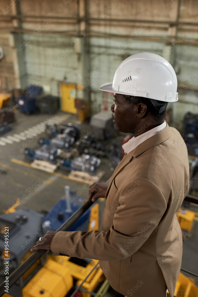 African engineer in work helmet standing on balcony on top floor and watching for the work in warehouse