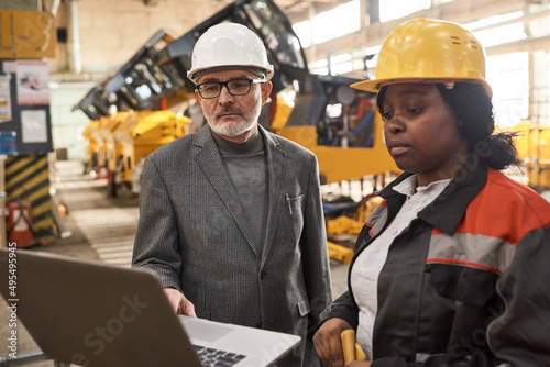 Senior engineer in work helmet using laptop to present new machines to worker who looking at monitor standing near