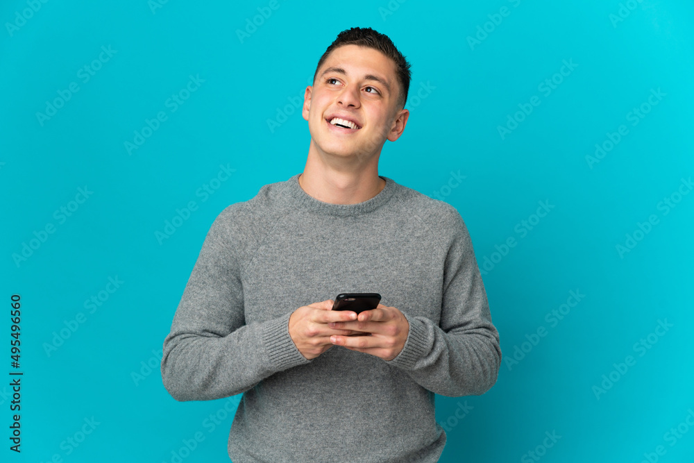 Young caucasian man isolated on blue background using mobile phone and looking up Young caucasian man isolated on blue background using mobile phone and looking up