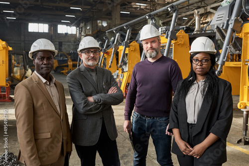 Portrait of multiracial business people in work helmets looking at camera while standing in factory with new equipment in the background