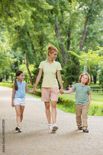 happy woman holding hands with kids while walking in park.