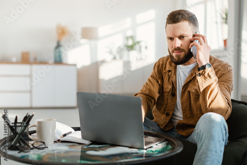 Serious Male Freelancer Talking On Phone Using Laptop At Home