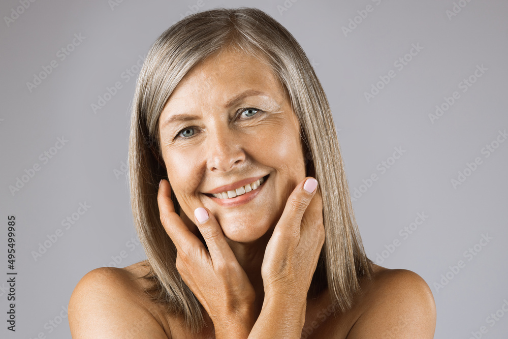 Portrait of caucasian aged woman with bare shoulders smiling on camera over grey background. Pretty lady having healthy smooth skin with natural wrinkles on face. Portrait of caucasian aged woman with bare shoulders smiling on camera over grey background. Pretty lady having healthy smooth skin with natural wrinkles on face.