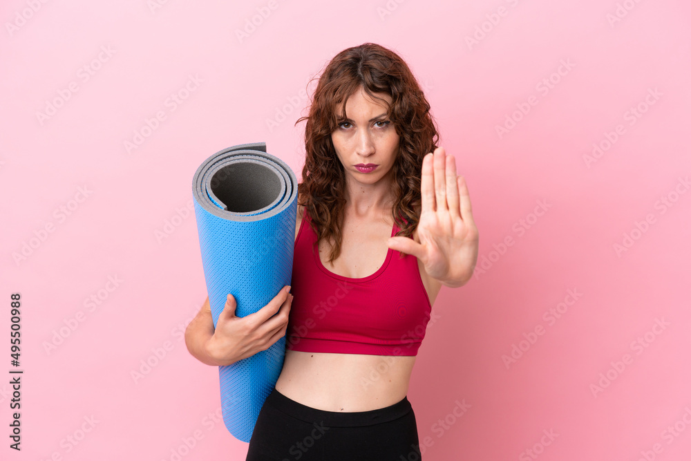 Young sport woman going to yoga classes while holding a mat isolated on pink background making stop gesture