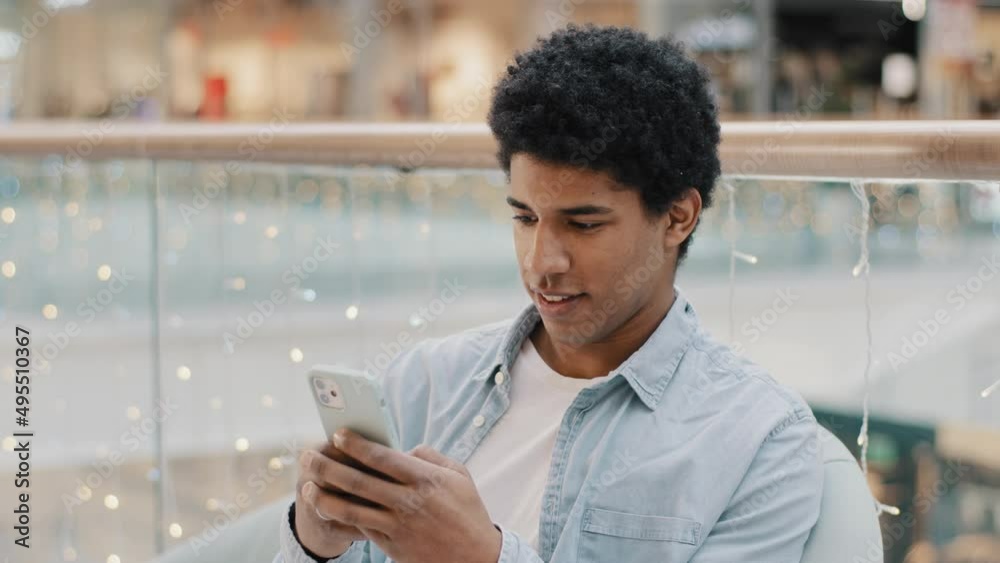 African focused millennial man sitting indoors swipes in phone screen mobile chat app online shopping play game with smartphone chatting browsing telephone free wi-fi user cellular chat social media 