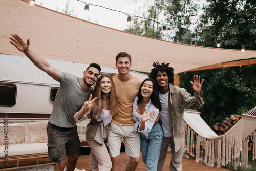 Portrait of cheery young international friends posing near camper van, smiling and waving at camera, having fun together