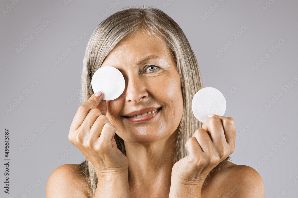 Portrait of mature caucasian woman holding white cosmetic sponges and smiling on camera over grey background. Natural beauty and skin care concept. Portrait of mature caucasian woman holding white cosmetic sponges and smiling on camera over grey background. Natural beauty and skin care concept.