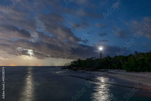 Near Fort Myers, Florida, the rising moon shines on San Carlos Bay as the Ybel Lighthouse shines on the beach at the tip of Sanibel Island while stars twinkle in the cloudy night sky.