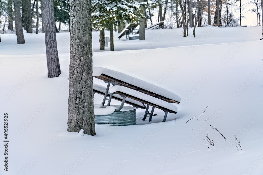 Snow covered picnic table leaned against a tree and fire pit in campground at Interlochen State Park, Michigan