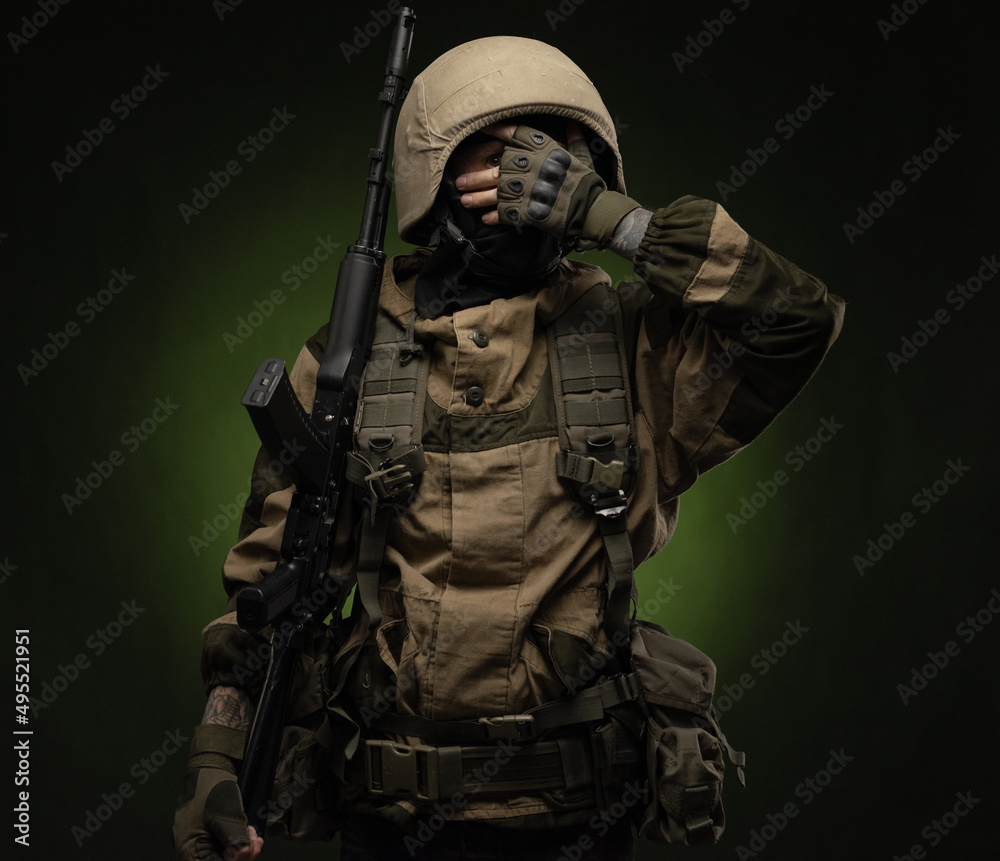 a soldier in military clothes and helmet, with a Kalashnikov rifle covers his face with his hand showing emotions on a dark background in the studio