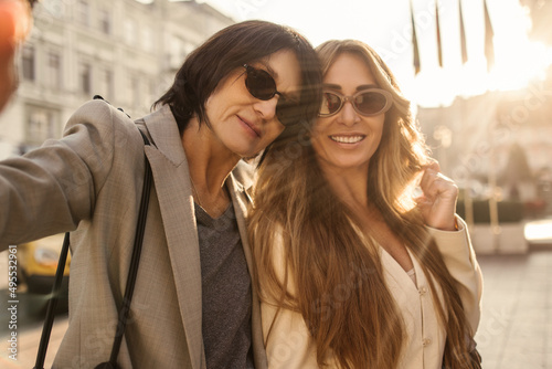 Two beautiful middle-aged caucasian women in sunglasses take selfie against backdrop of sunny city. Models in good mood spend time together on weekends.
