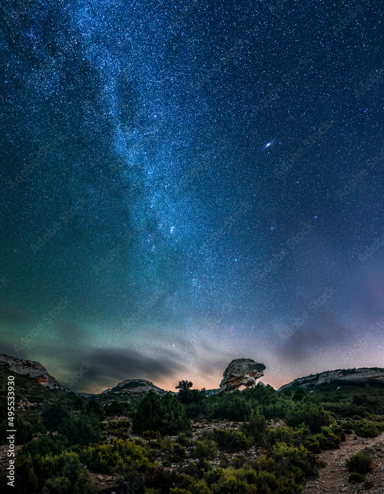Panoramic view of the Milky Way and the Andromeda galaxy over a rock arch shaped like a turtle. Night landscape