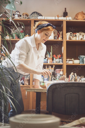 A young active female artist, with red hair, in a white shirt and jeans with a bandage on her head, paints standing at the table photo