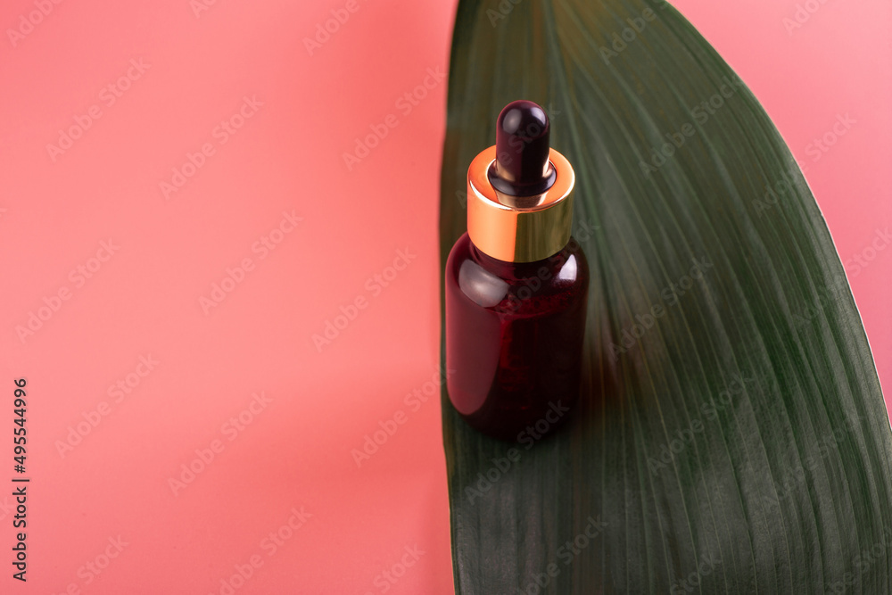 Obraz premium glass brown bottle with pipette with essential oil on a pink background with green tropical leaves top view. Aromatic cosmetic product for skin and hair care Moisturizing beauty product with organic