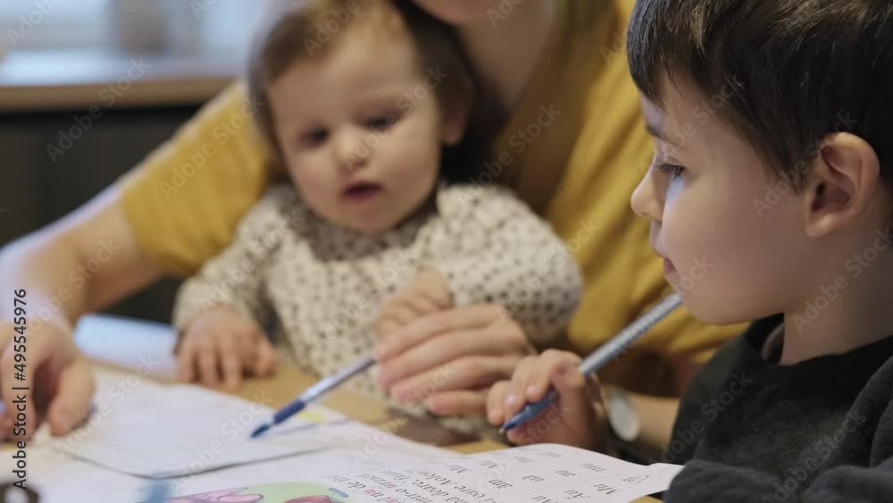 Boy doing homework with the help of his loving parent. Mum helping her boy with homework while holding baby daughter in arms. Studying, doing homework. Boy doing homework with the help of his loving parent. Mum helping her boy with homework while holding baby daughter in arms. Studying, doing homework.